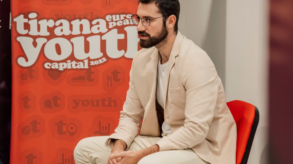 Man sitting at the Tirana Youth Capital 2022 event, wearing a blazer and eyeglasses.